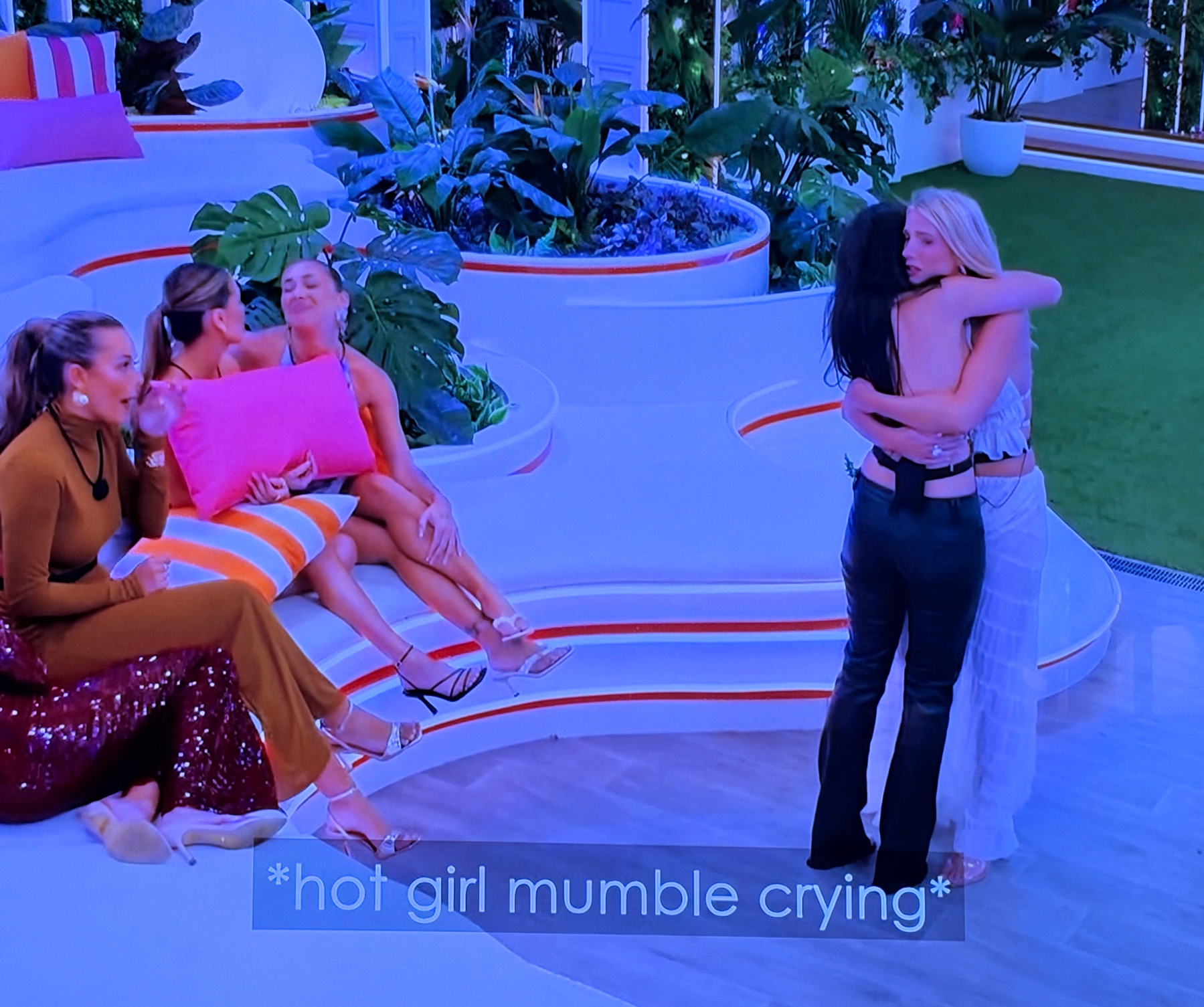 Screenshot from “love island Australia”. To the left of the frame several female contestants sit on an outdoor sofa. They’re wearing evening wear and chatting. To the right, two additional female contestants, also in evening wear, are standing and hugging. The caption is from the program’s own closed captioning. It reads “hot girl mumble crying”.&10;